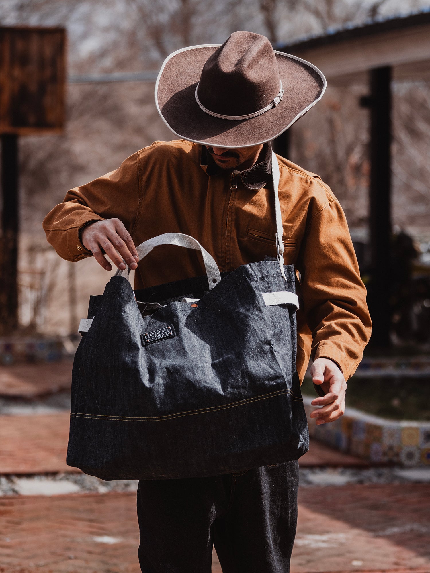 Person wearing a brown hat and jacket, holding a dark blue bag outdoors.