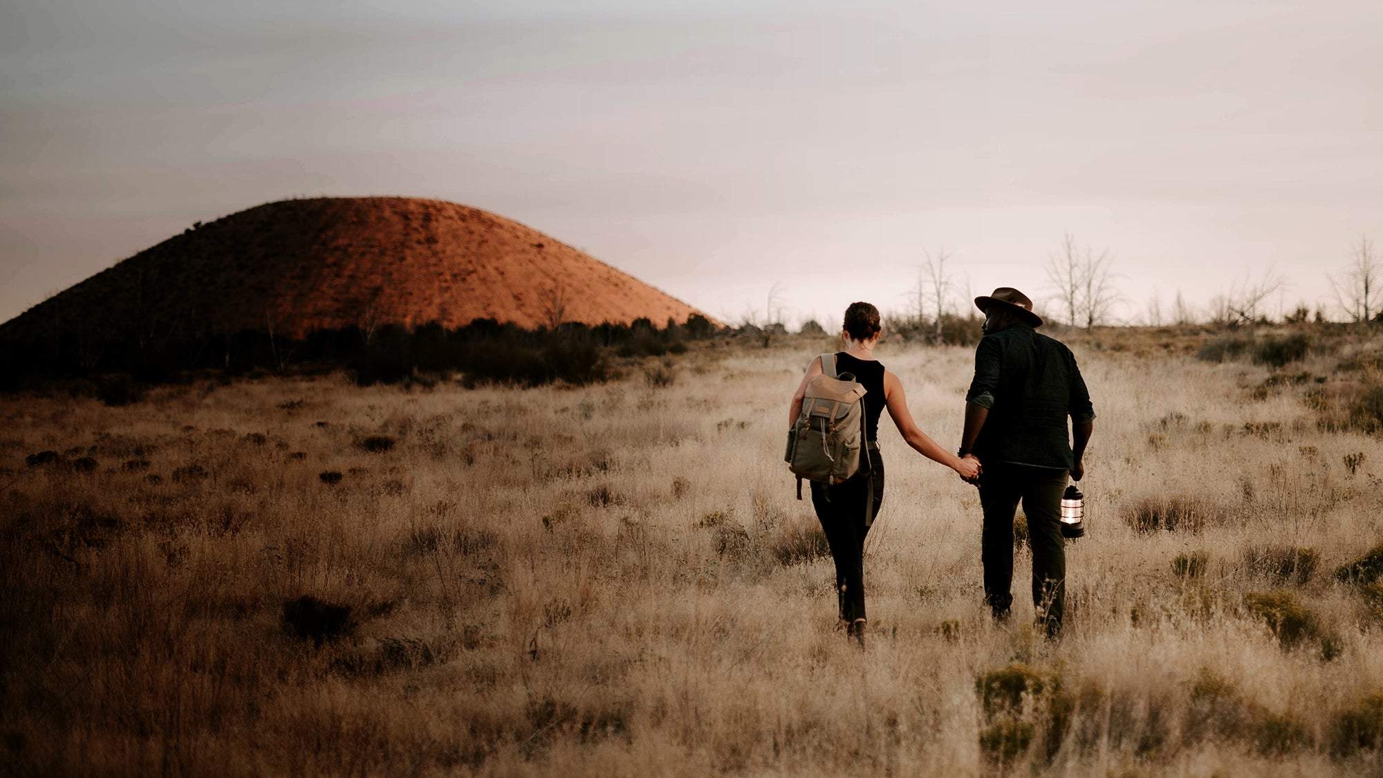 Two people walking through a dry field holding hands, one carrying a Barebones Forest Lantern.