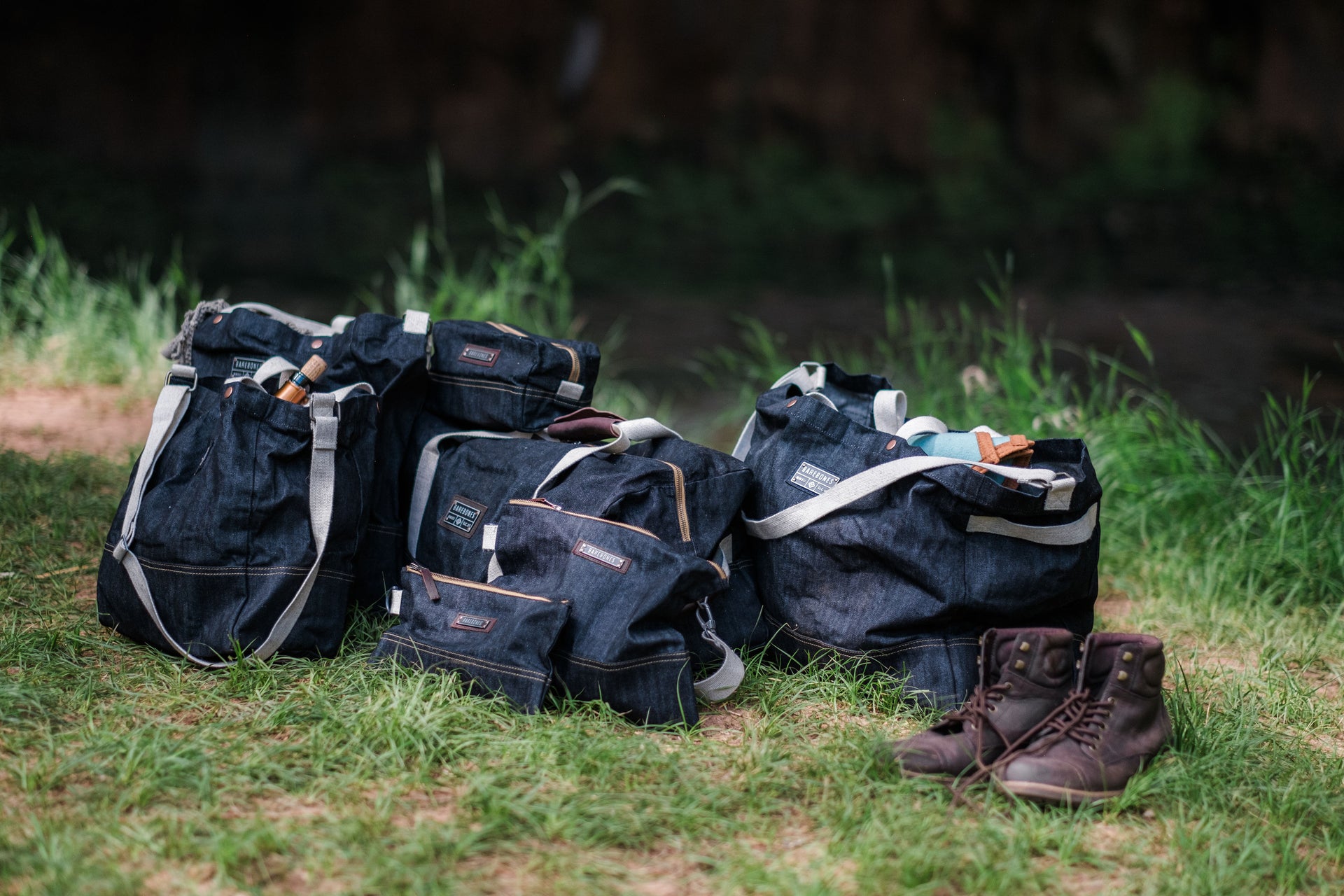 A person uses a durable waxed canvas bag to carry firewood to an open fire. They unload the logs and place them into the flames before reaching into another bag for cooking tools. The sturdy bags, designed for outdoor use, hold essentials for open-fire cooking, making transport and organization easy in the rustic setting.